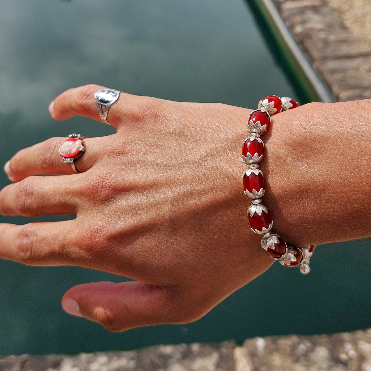 A close-up of a person's hand and wrist wearing a red and silver beaded bracelet, with matching silver rings, posed in front of a pond.