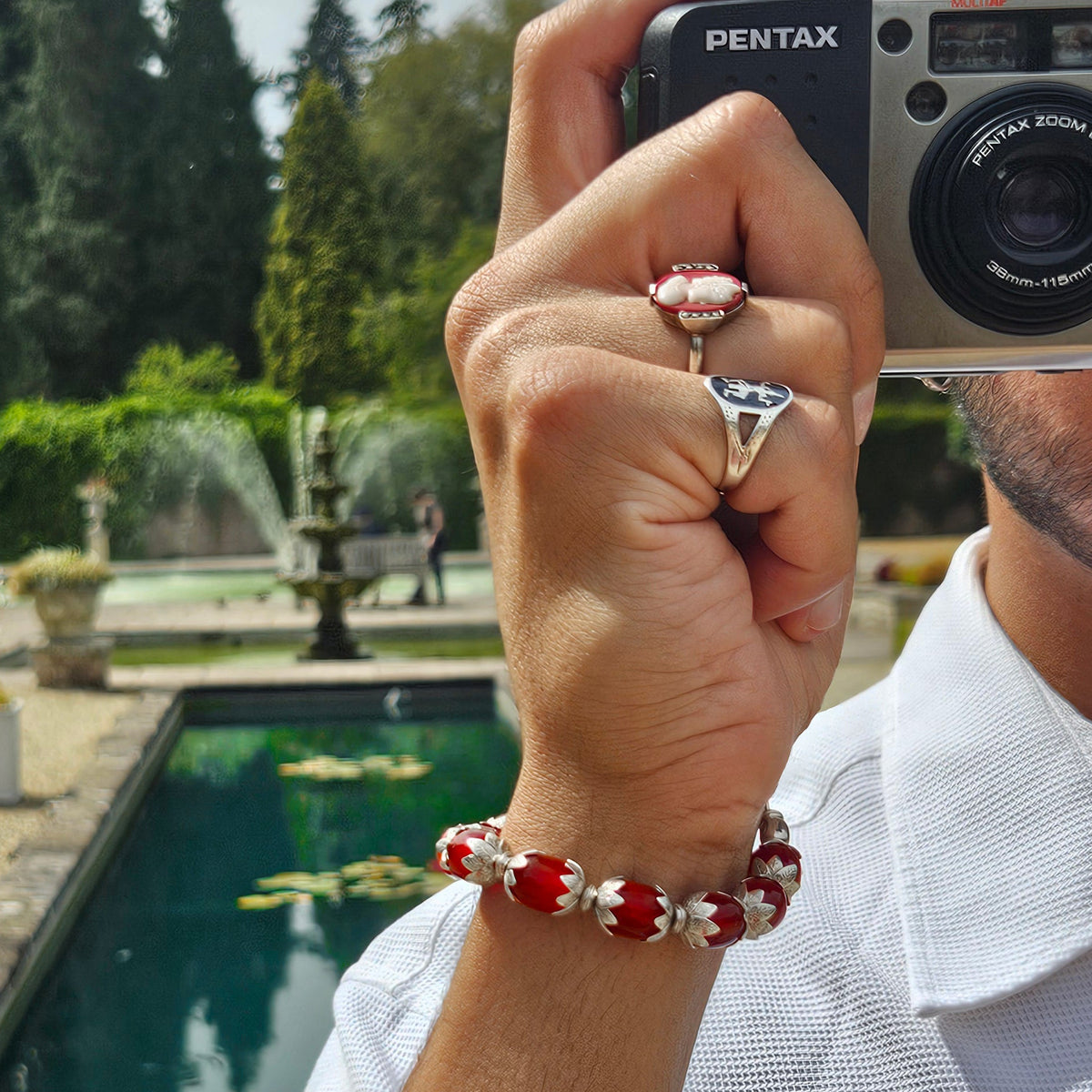 A close-up lifestyle shot of a man wearing a red and white beaded bracelet on his wrist, holding a silver Pentax camera to take a mirror selfie in a garden setting.