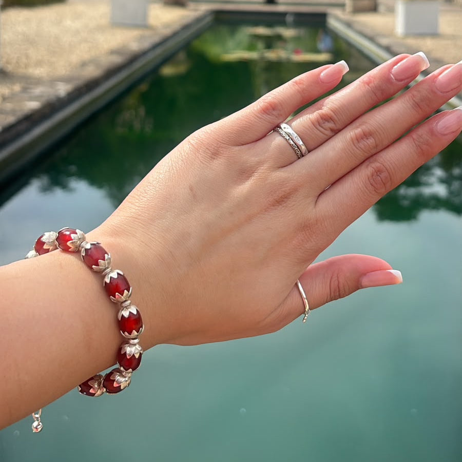 A close-up of a person's hand wearing a red and silver beaded bracelet and a silver ring, set against a blurred background of a garden pool and trees.